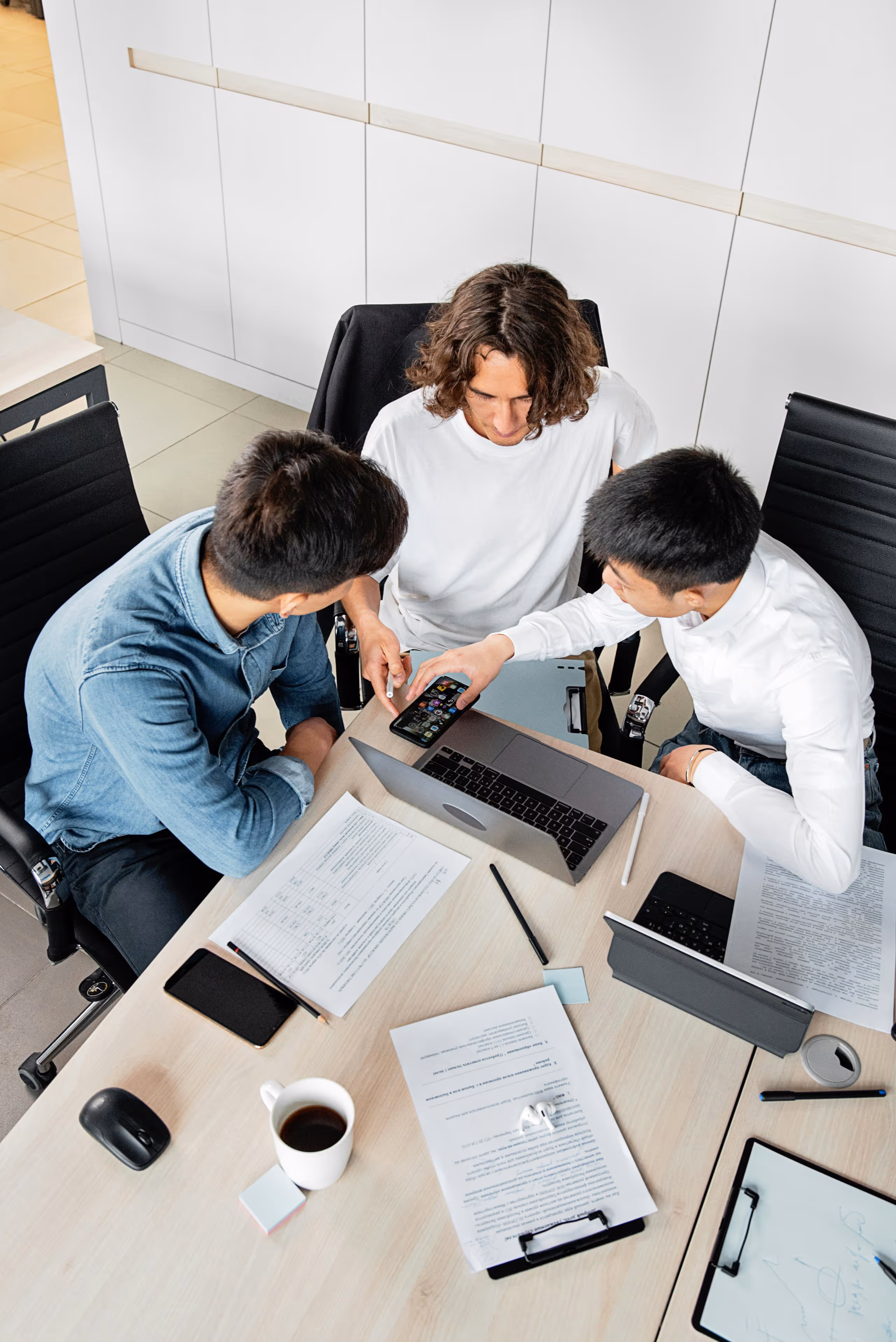 Three people sitting around an office table, looking at a smartphone, with a laptop, documents, and a coffee cup on the table.