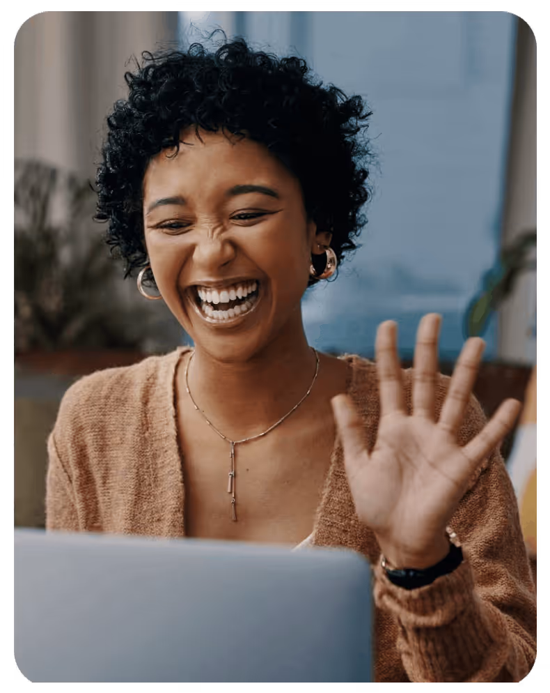 Smiling woman with curly hair waving her hand while looking at a laptop screen.