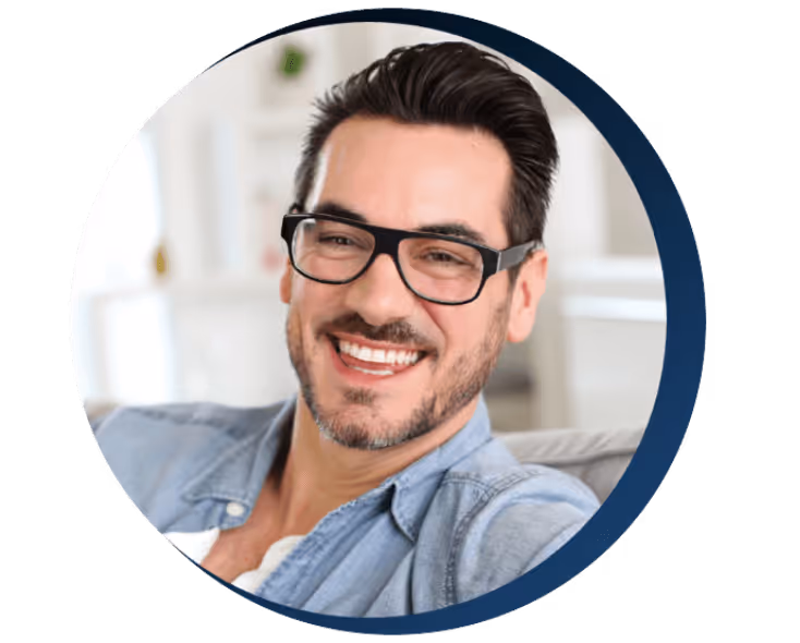 Smiling man with dark hair, glasses, and a beard wearing a light denim shirt indoors.
