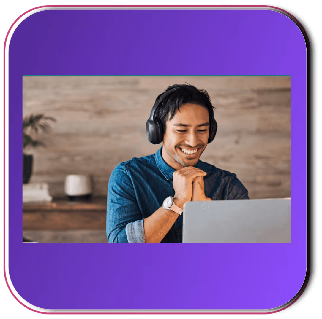 Smiling man wearing headphones and a denim shirt, sitting indoors and looking at a laptop screen.