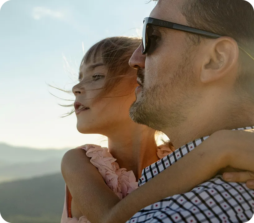 Man wearing sunglasses holding a young girl with a pink dress outdoors at sunset.