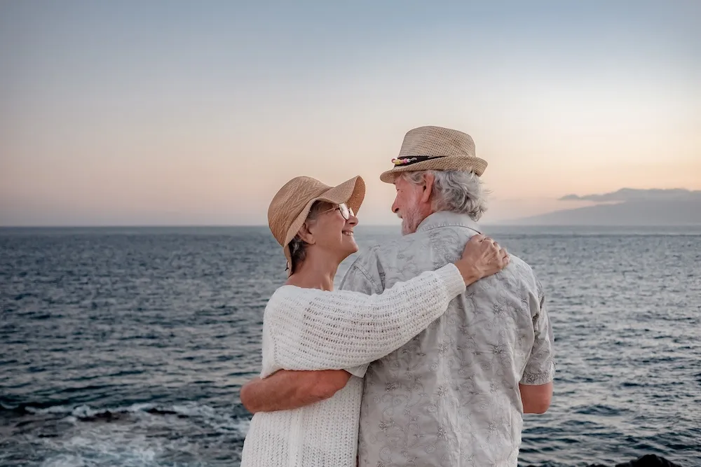Senior couple wearing hats embracing and smiling by the ocean at sunset.