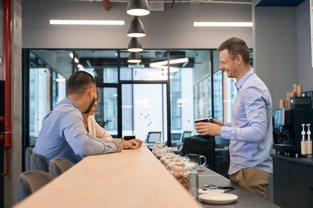 Two office workers talking with a barista holding a cup in a modern office coffee area.