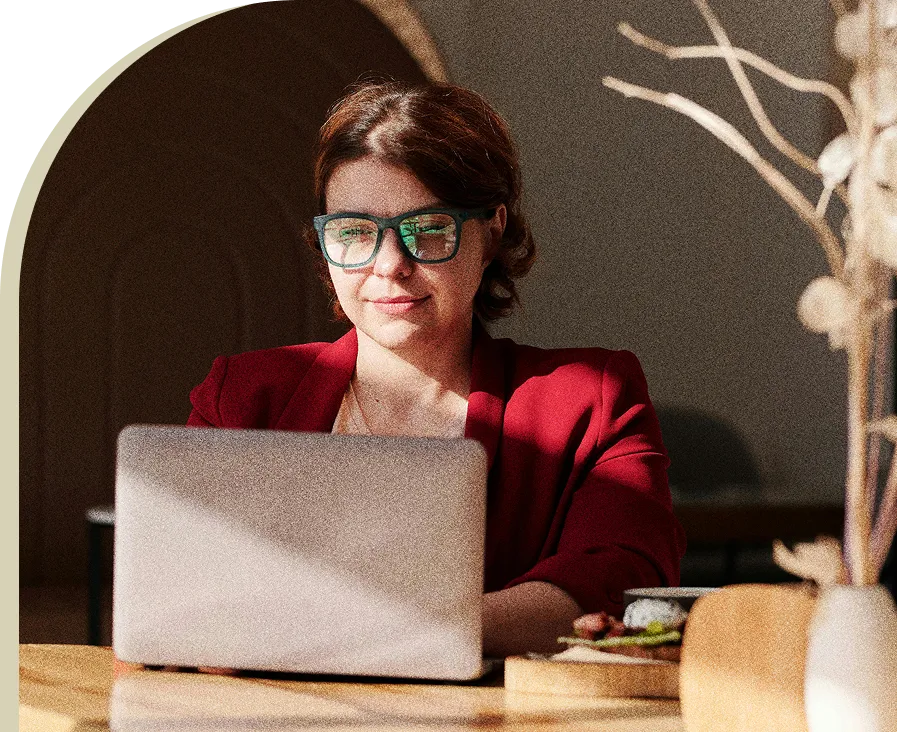 Woman with glasses and a red blazer working on a laptop at a wooden table in natural light.