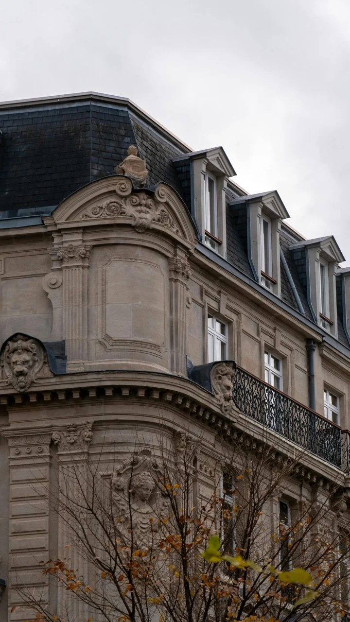 Ornate historic building corner with sculpted stone details, wrought iron balcony, and dormer windows under a cloudy sky.