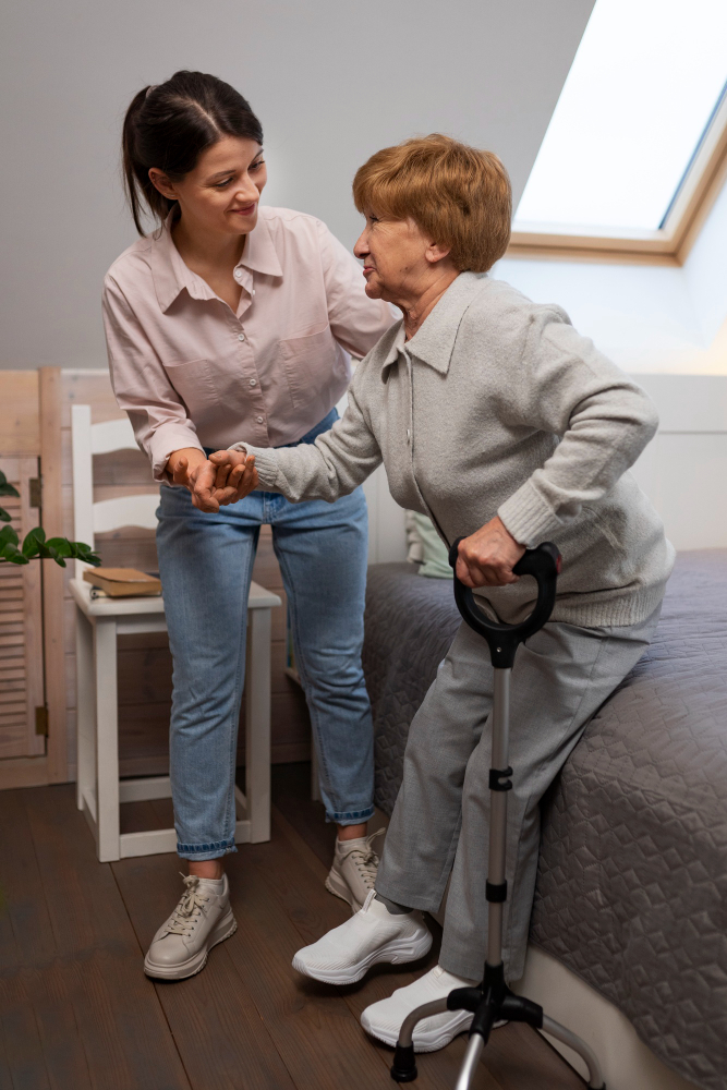 Caregiver helping an older woman stand beside a bed with a walking cane