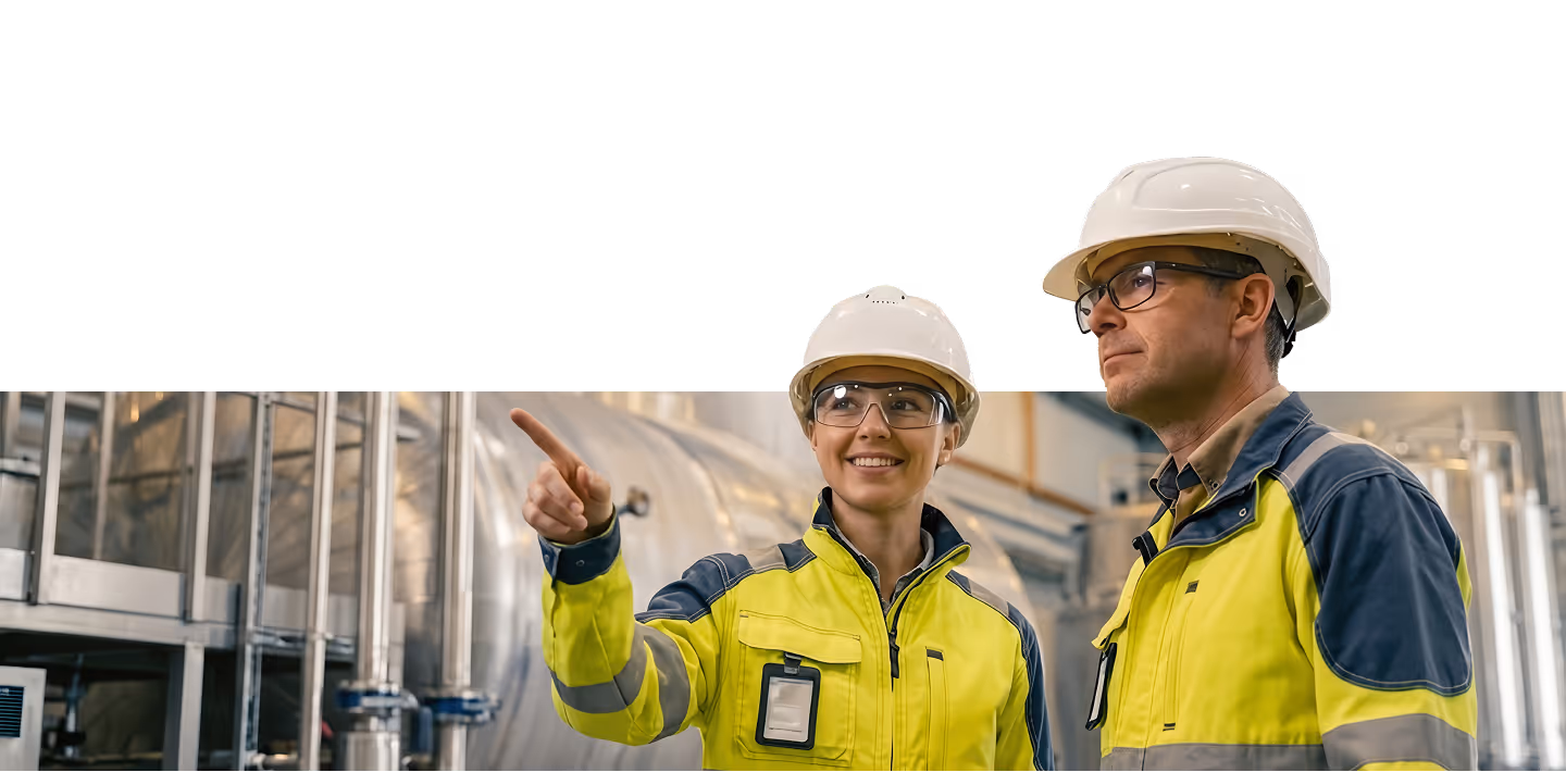 Two industrial workers wearing white hard hats and yellow safety jackets, one pointing and smiling while the other looks ahead in a factory setting.