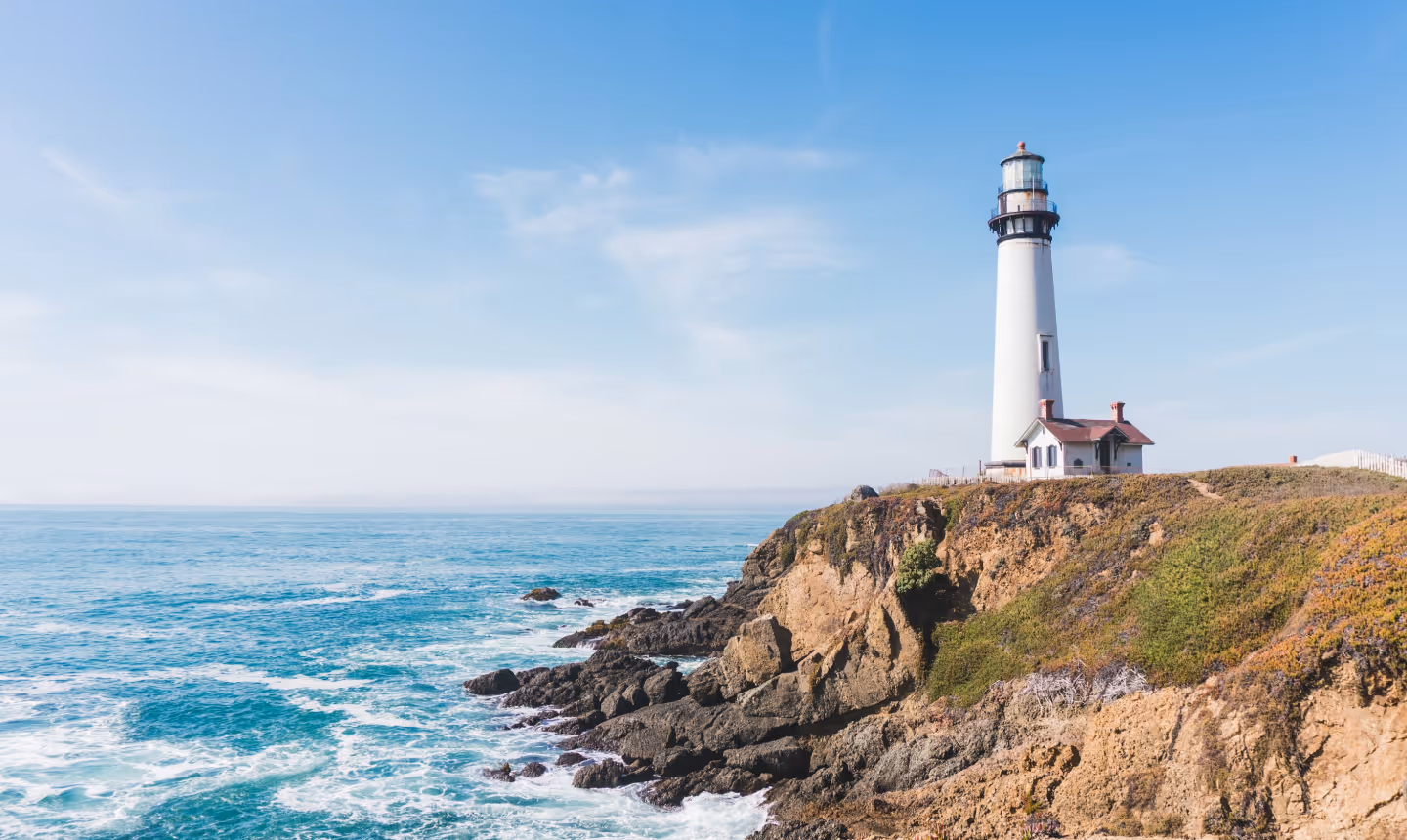 White lighthouse on a rocky cliff overlooking the ocean under a clear blue sky.