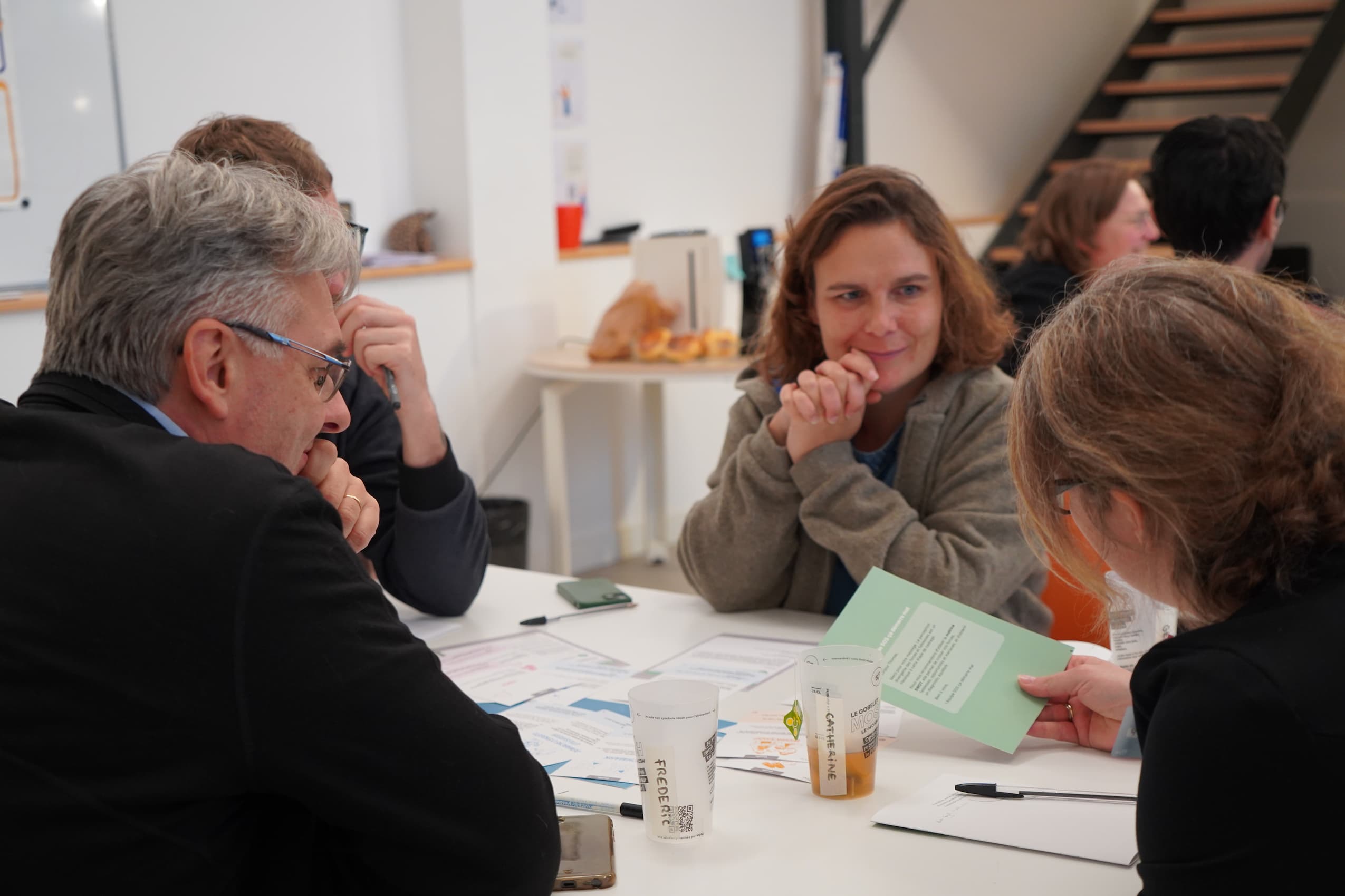 Quatre personnes assises autour d'une table blanche discutant et examinant des documents lors d'une réunion informelle avec des gobelets en plastique.