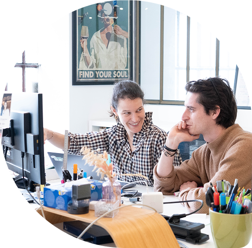 Deux collègues souriants discutant devant un ordinateur dans un bureau lumineux avec une affiche « Find Your Soul » en arrière-plan.