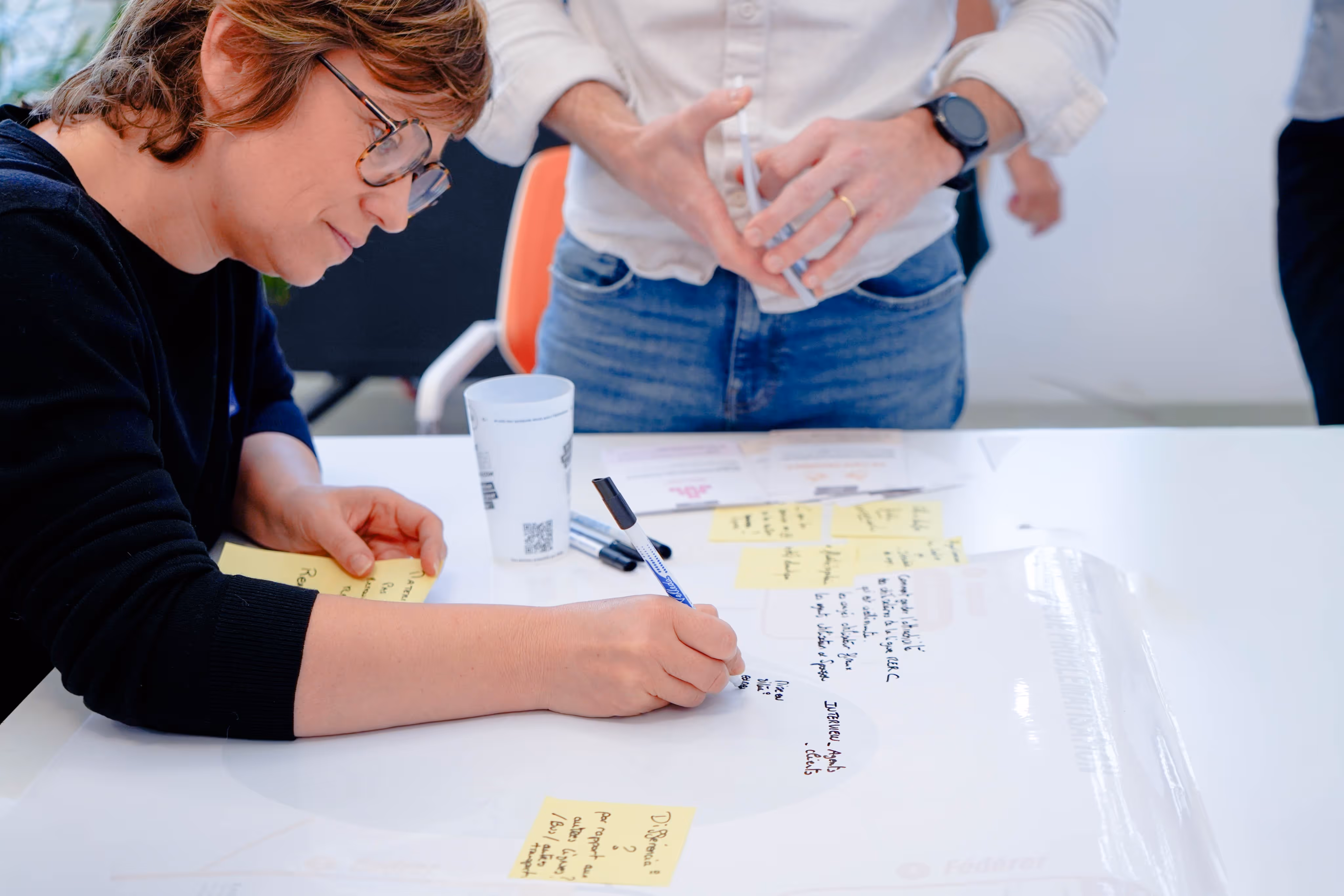 Person wearing glasses writing on a large white sheet with sticky notes and handwritten notes during a group discussion.