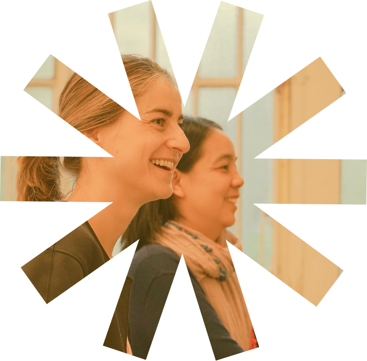 Deux femmes souriantes assises côte à côte devant une fenêtre avec rideaux translucides.