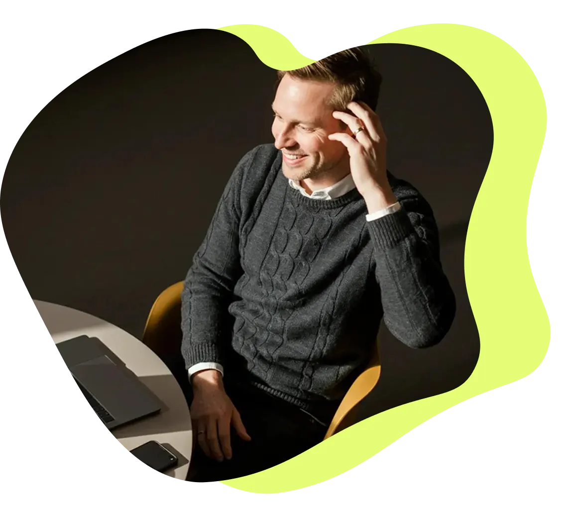 Leende man i grå stickad tröja sitter vid ett bord med en bärbar dator och en mobiltelefon.