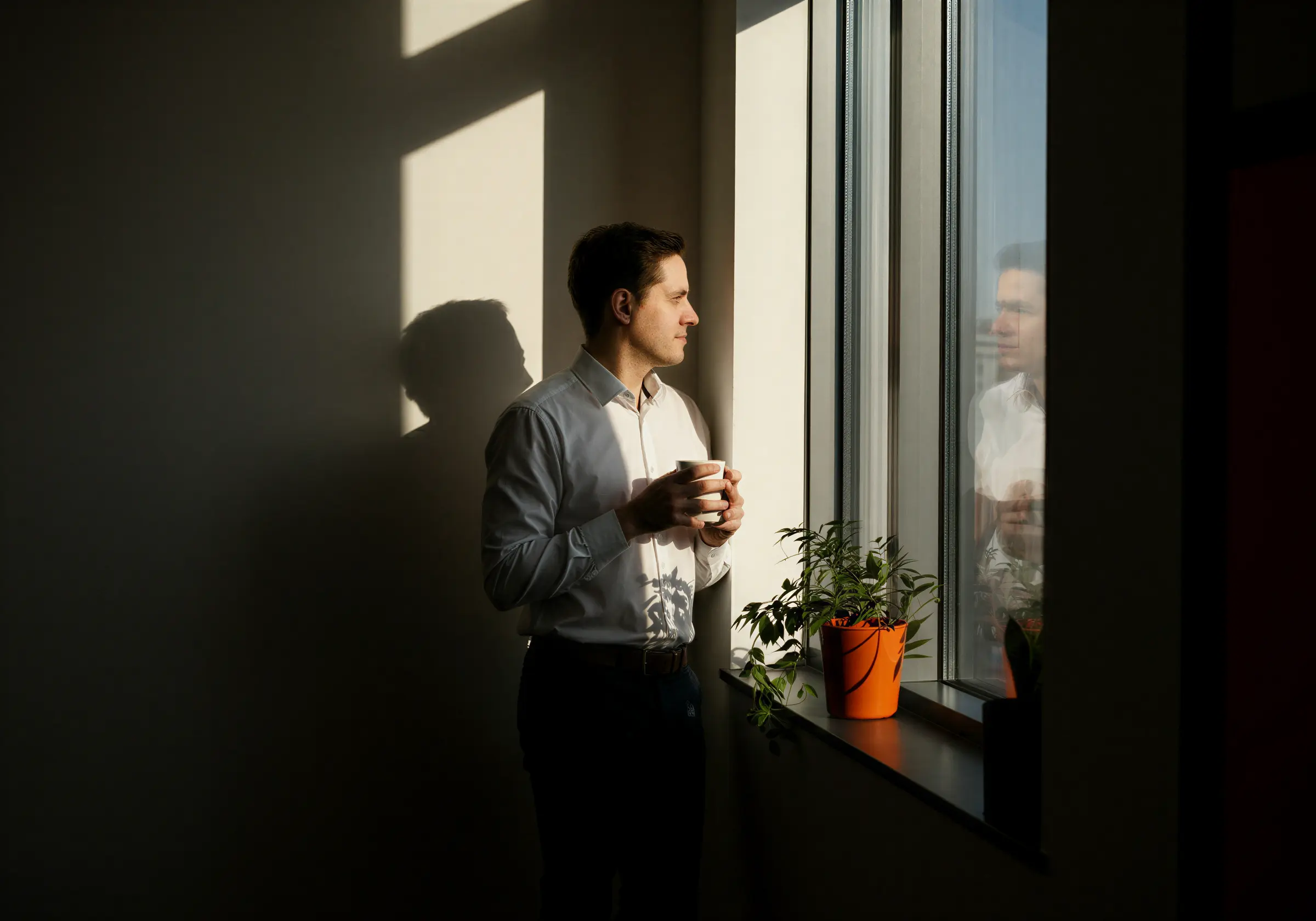 Man i vit skjorta som håller en kopp medan han ser ut genom ett fönster med en växt i en orange kruka på fönsterbrädan.