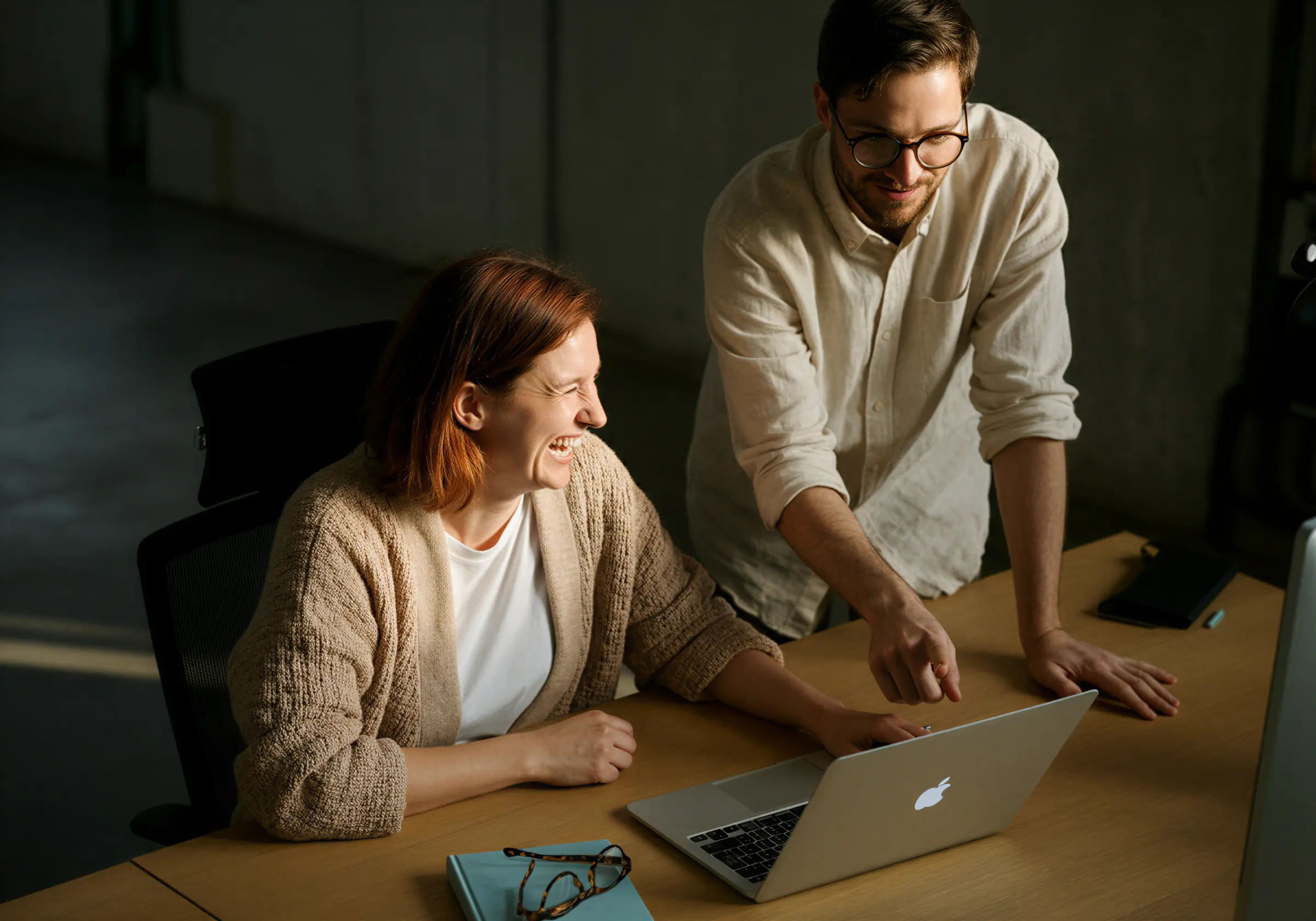 Två kollegor samarbetar och skrattar medan de tittar på en bärbar dator vid ett skrivbord.
