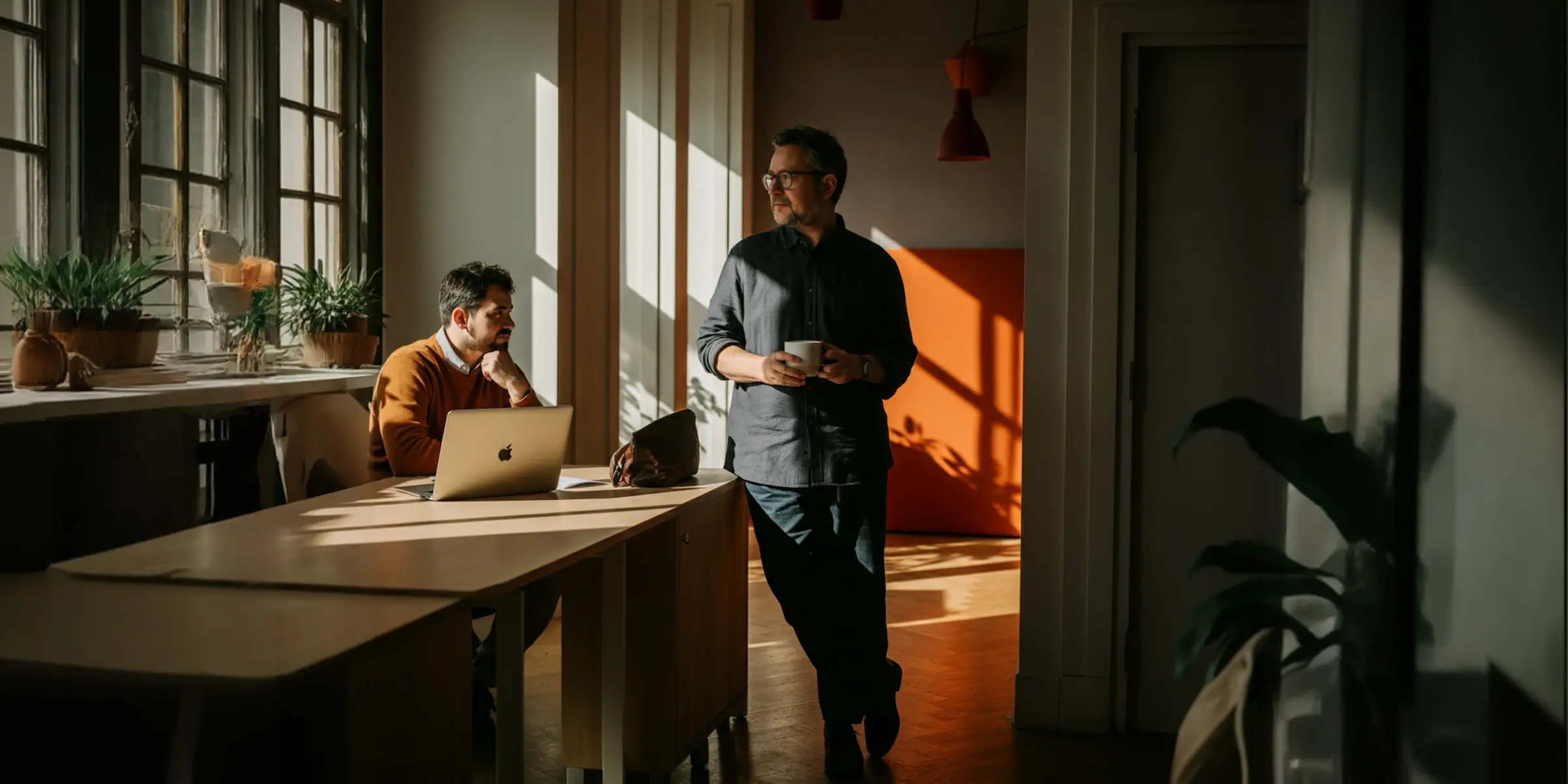 Två män i ett solbelyst kontor, den ena sitter vid ett skrivbord med en laptop, den andra står och håller en kopp.