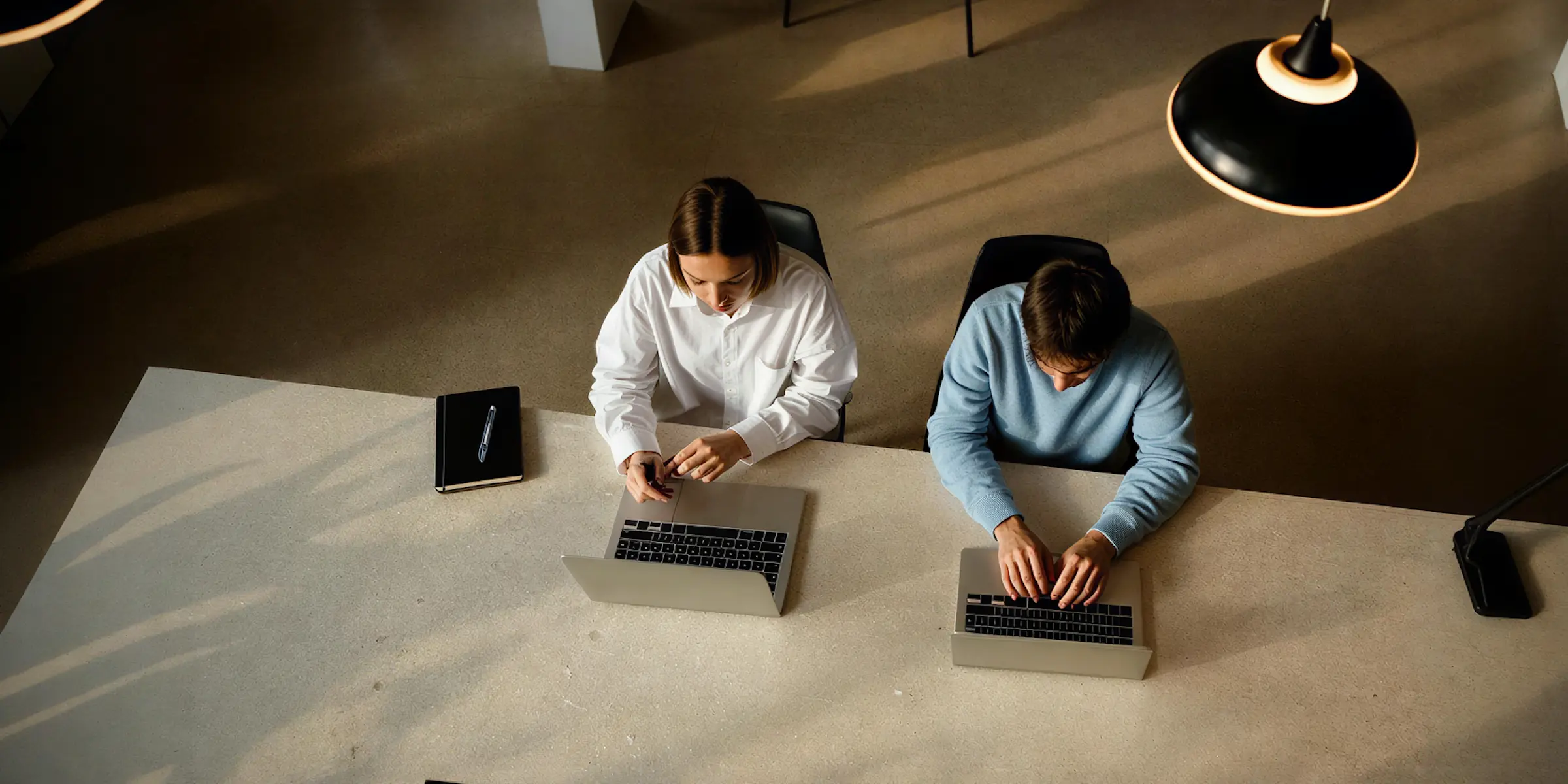 Två personer som arbetar med bärbara datorer vid ett stort bord i ett väl upplyst kontorsutrymme.