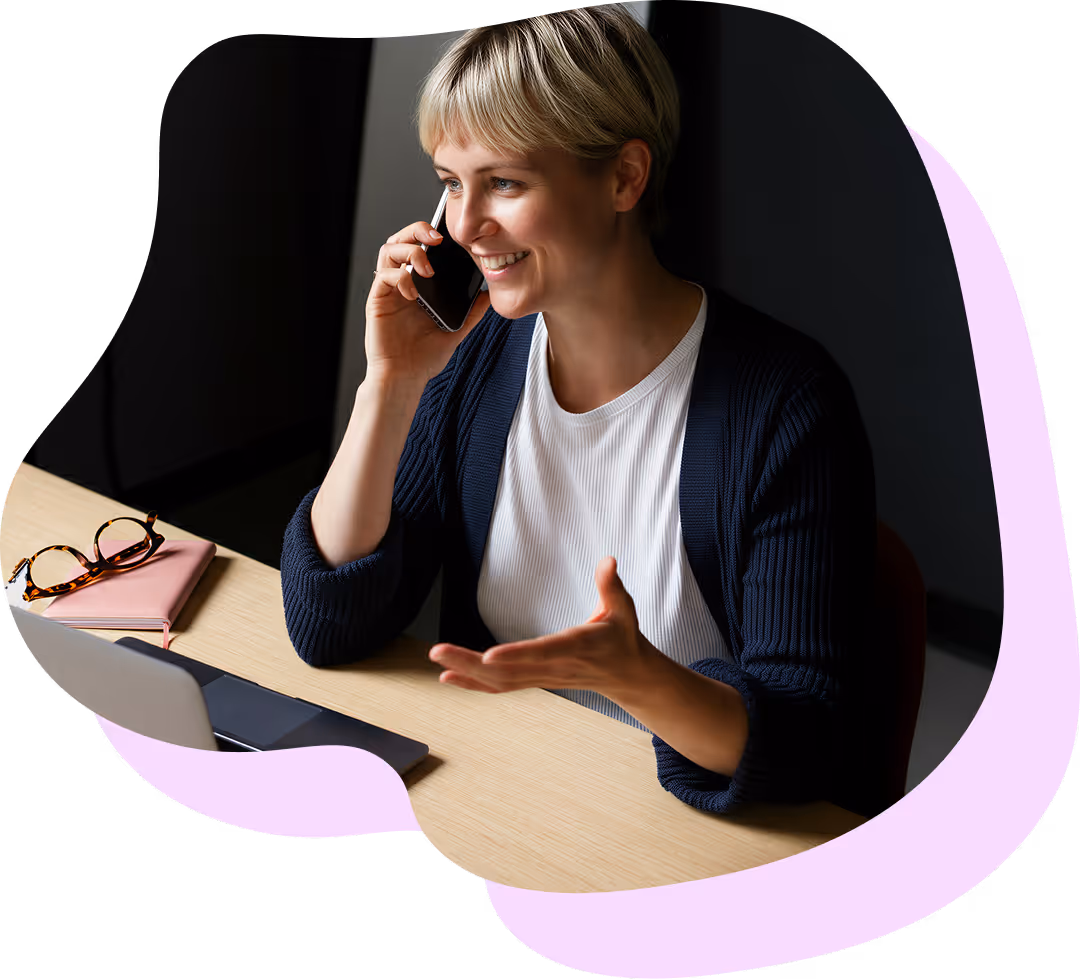 Leende kvinna med kort blont hår som pratar i mobiltelefon vid ett skrivbord med en öppen bärbar dator och ett par glasögon på en anteckningsbok.