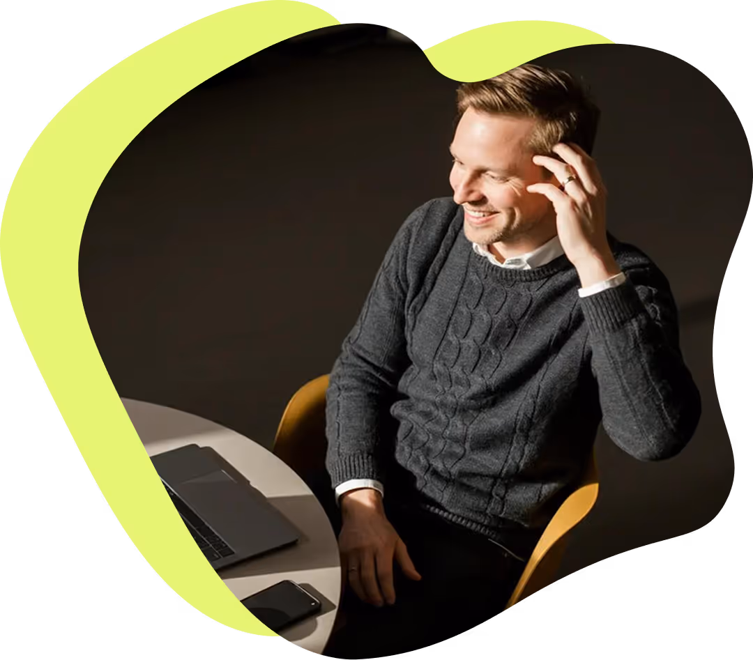 Glad man i mörk tröja sitter på en stol vid ett bord med en bärbar dator och en mobiltelefon.