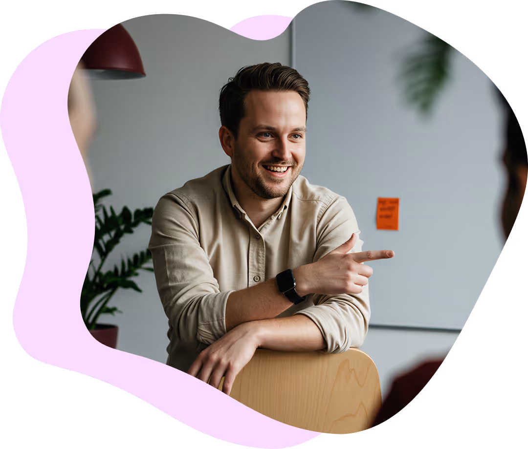 Leende man i beige skjorta lutar sig mot en stol och pekar åt höger i ett kontorsrum med växt i bakgrunden.