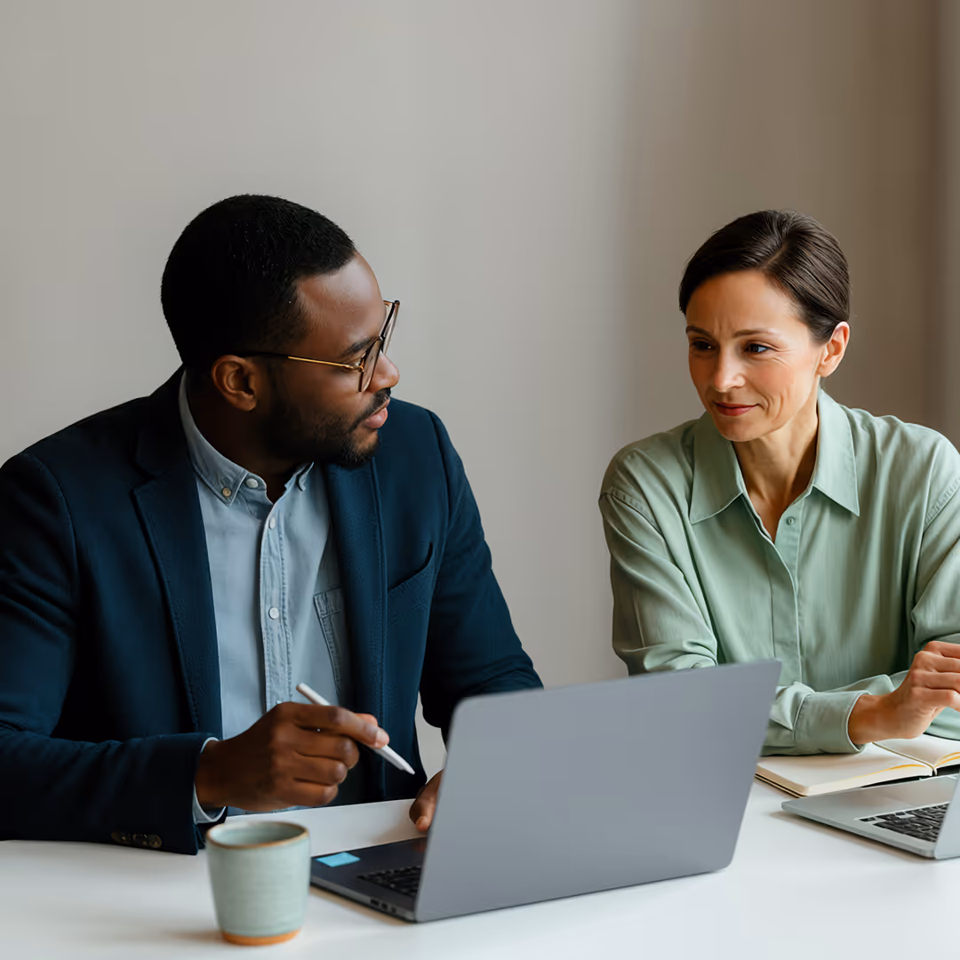 Två kollegor i samtal vid ett bord med bärbara datorer, en man visar något på sin skärm med en penna medan kvinnan lyssnar.