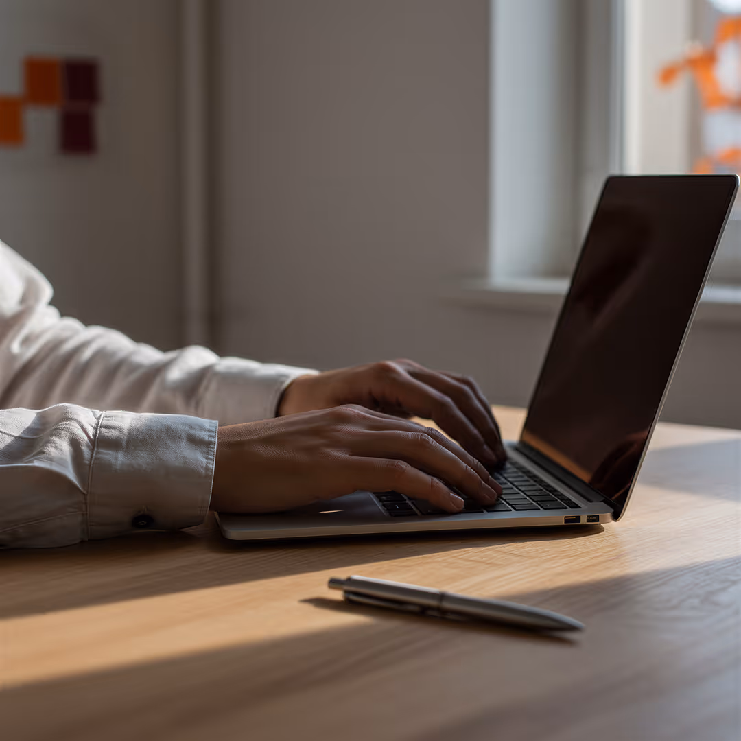 En person som skriver på en bärbar dator på ett träbord med en penna framför.
