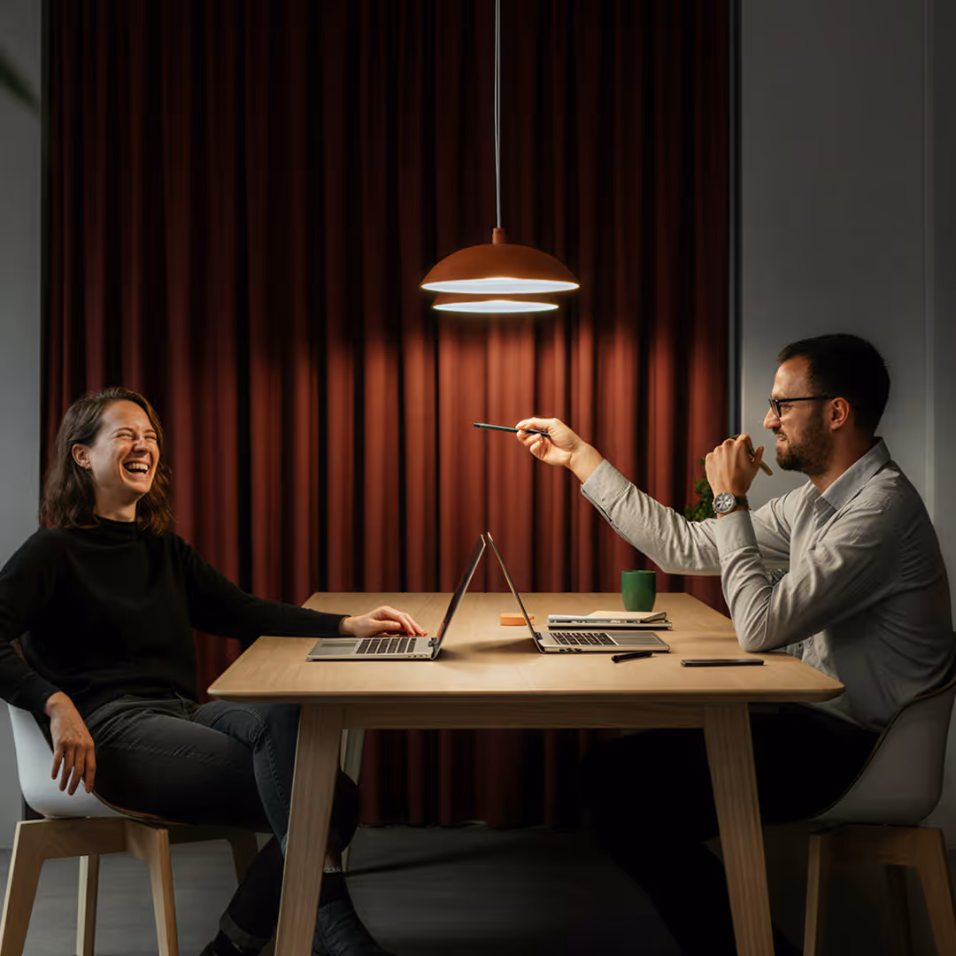 Två kollegor sitter vid ett bord med bärbara datorer och har en livlig konversation under en taklampa.