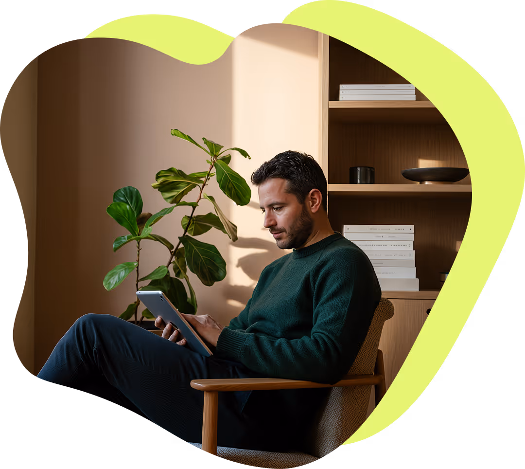 Man med grön tröja sitter i en stol och använder en surfplatta i ett rum med en stor grön växt och bokhylla i bakgrunden.
