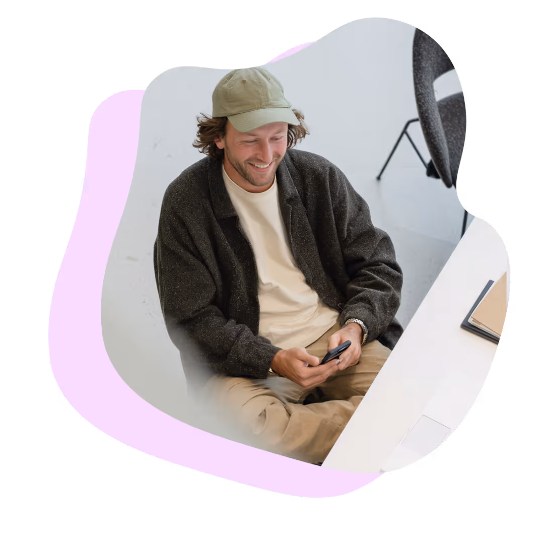 En leende man i keps och mörk jacka sitter och tittar på sin mobiltelefon vid ett skrivbord.