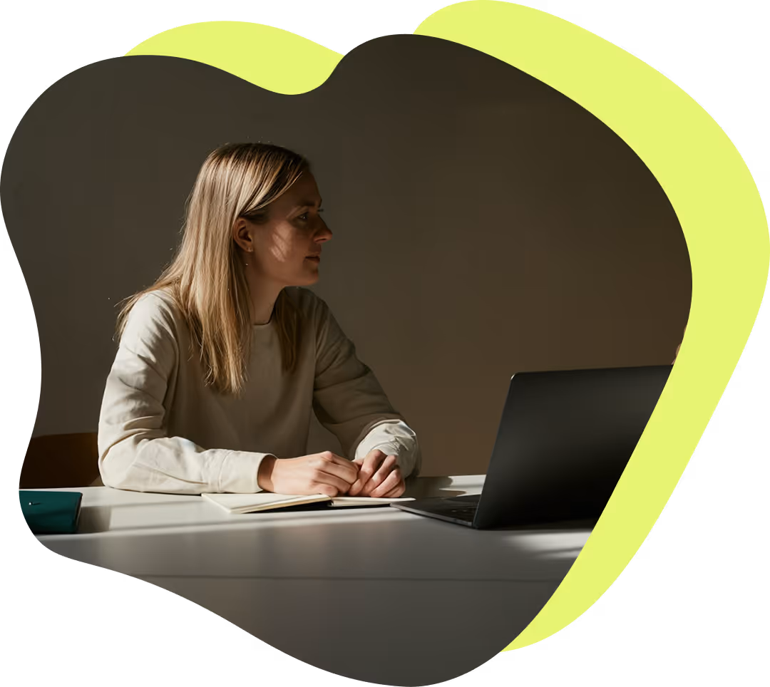 En kvinna med långt blont hår sitter vid ett skrivbord och tittar mot en öppen bärbar dator med en anteckningsbok framför sig.