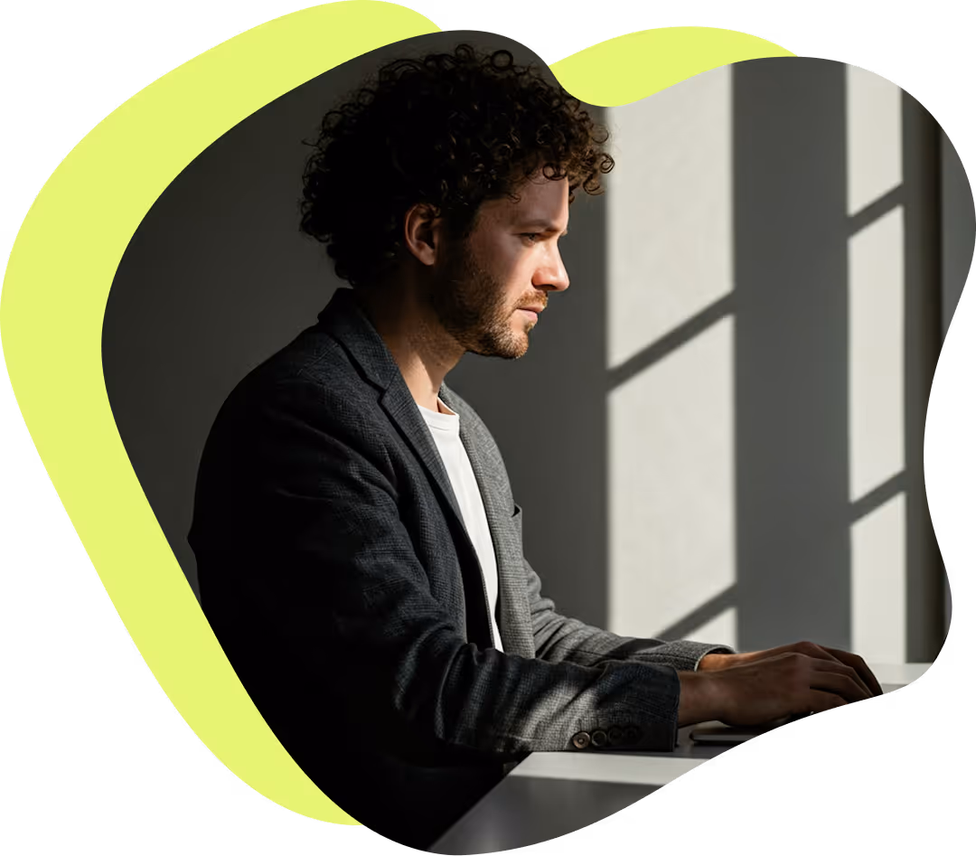 Man med lockigt hår och kavaj arbetar fokuserat vid ett bord med solljus som kastar skuggor från ett fönster.