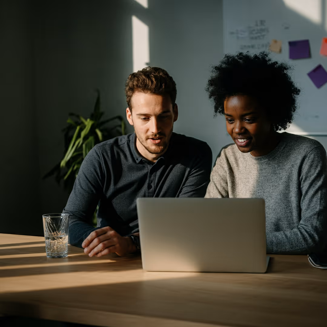 Två personer samarbetar och tittar på en laptop vid ett bord i ett solbelyst rum.