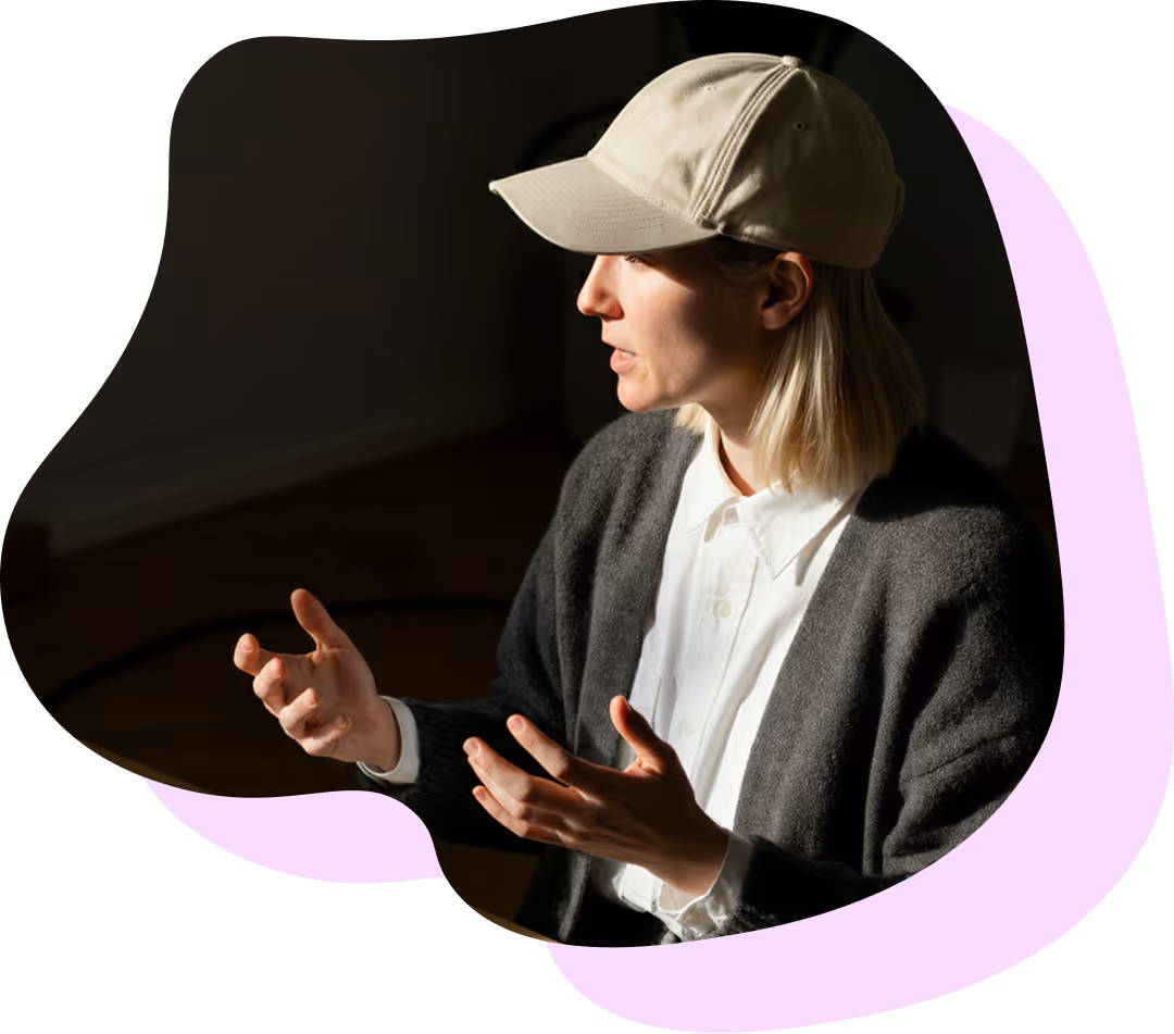 Kvinna med beige keps och grå tröja som talar med händerna framför en mörk bakgrund.