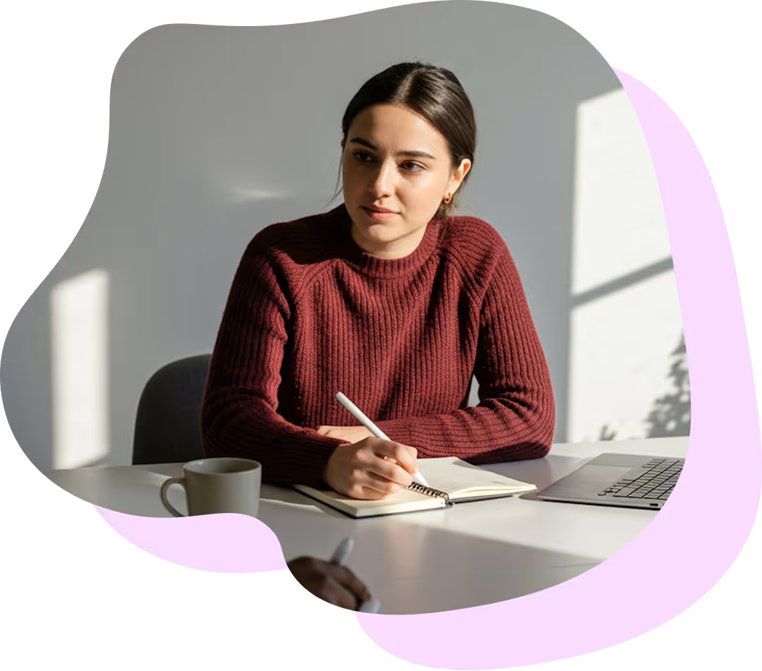 Ung kvinna i vinröd tröja som skriver i anteckningsbok vid ett bord med en kaffemugg och en laptop.