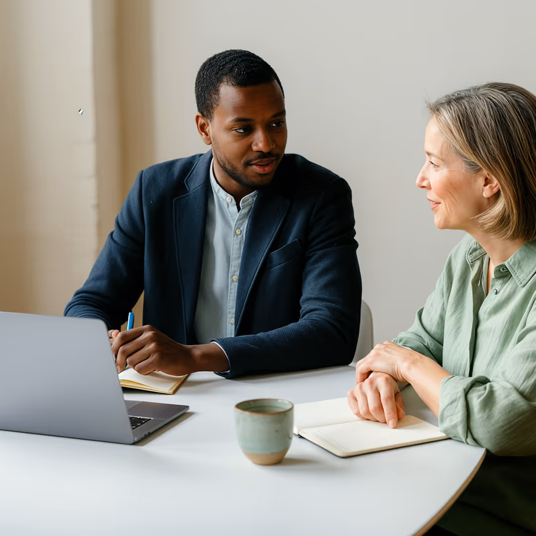 Två personer, en man och en kvinna, har ett professionellt samtal vid ett bord med en bärbar dator och anteckningsböcker.