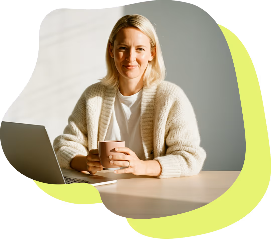 Leende kvinna med ljus tröja håller en kopp bredvid en bärbar dator vid ett bord i solljus.