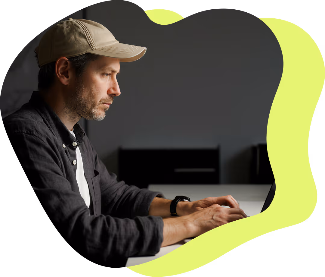 Man med keps och mörk skjorta arbetar koncentrerat vid en bärbar dator vid ett bord.