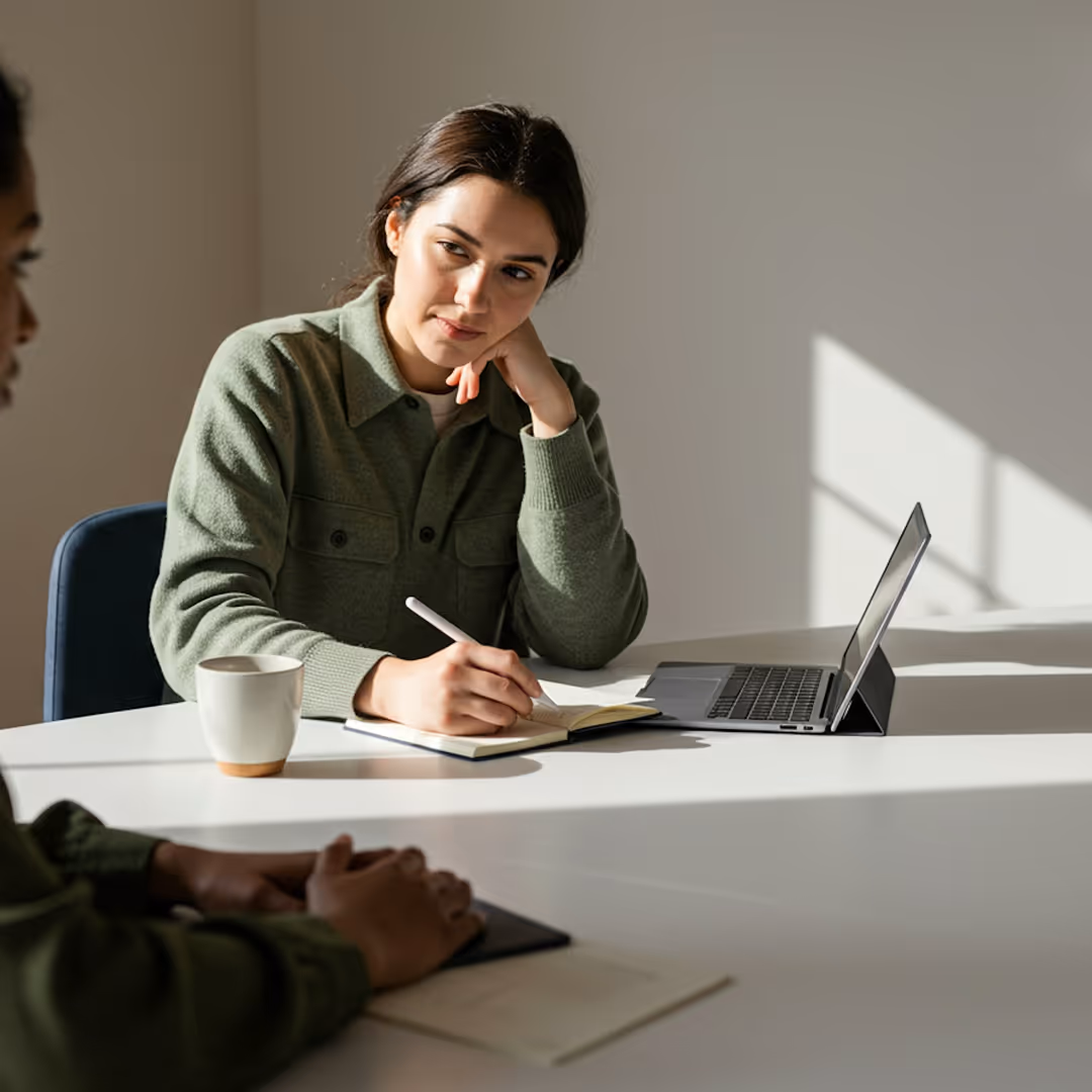 Kvinna som noterar i en anteckningsbok vid ett bord med en bärbar dator och en mugg under ett möte.