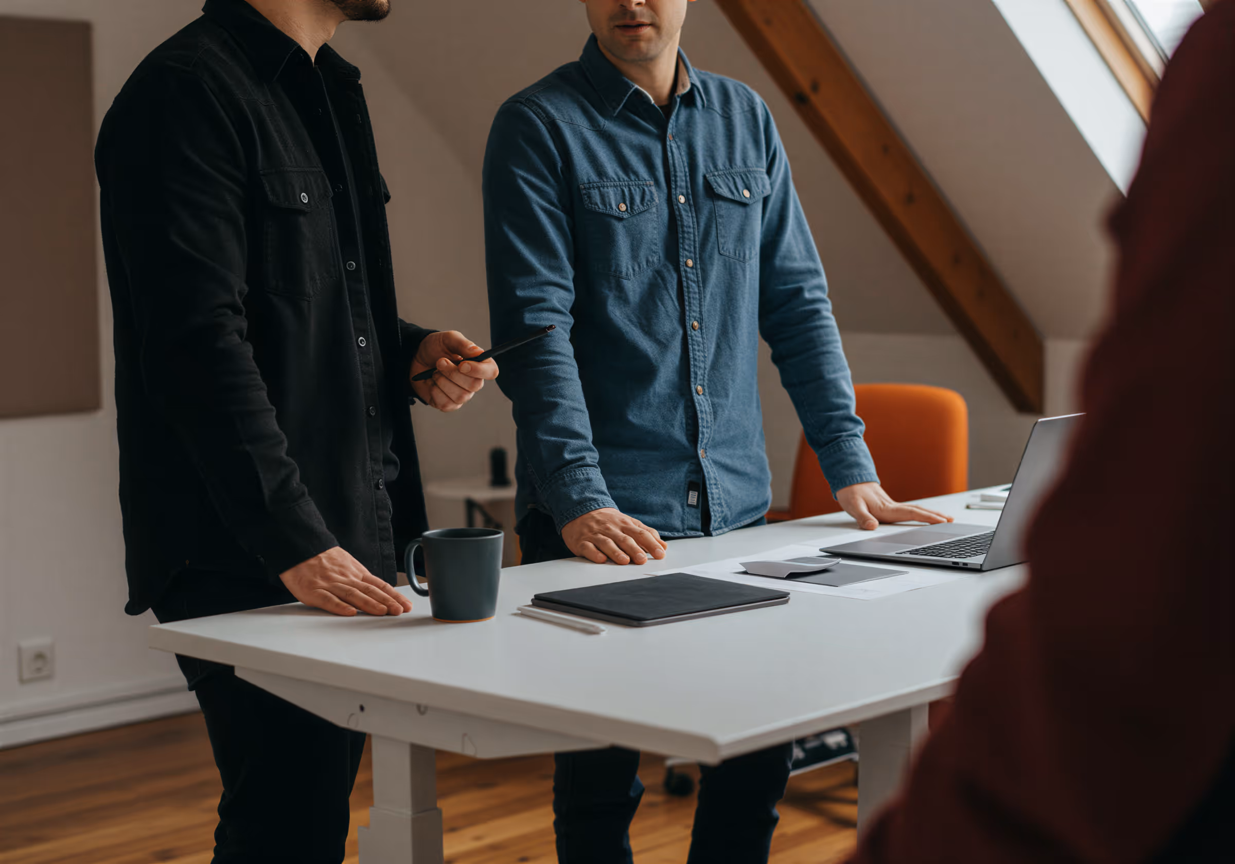 Två män som står och diskuterar vid ett vitt skrivbord med en bärbar dator, anteckningsbok och kaffemugg i ett kontorsrum med snedtak.