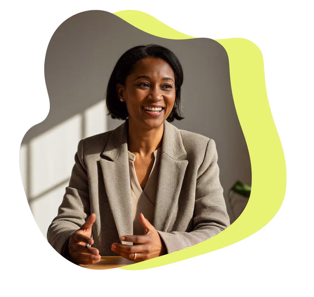 Leende kvinna i kavaj gestikulerar medan hon sitter vid ett bord med naturligt ljus i bakgrunden.