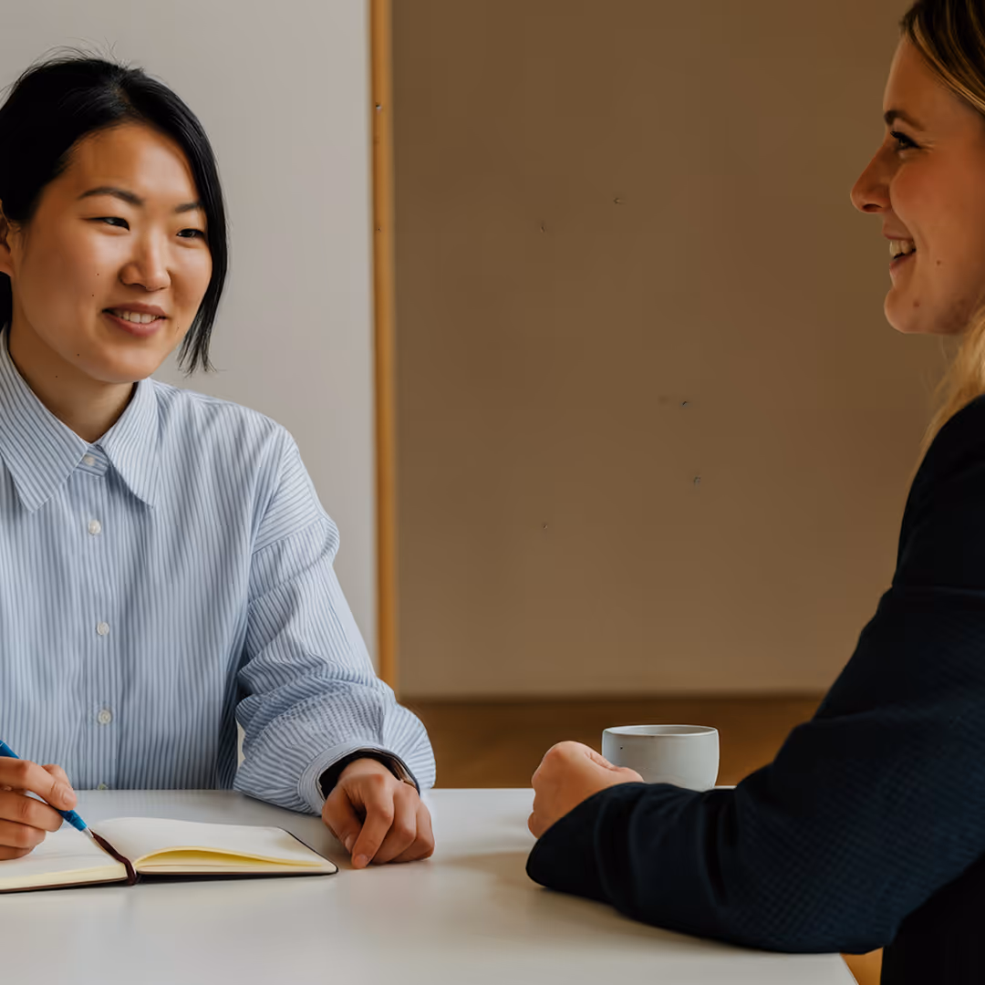 Två kvinnor i professionell miljö samtalar över ett bord, en antecknar i en bok medan den andra håller en kopp.