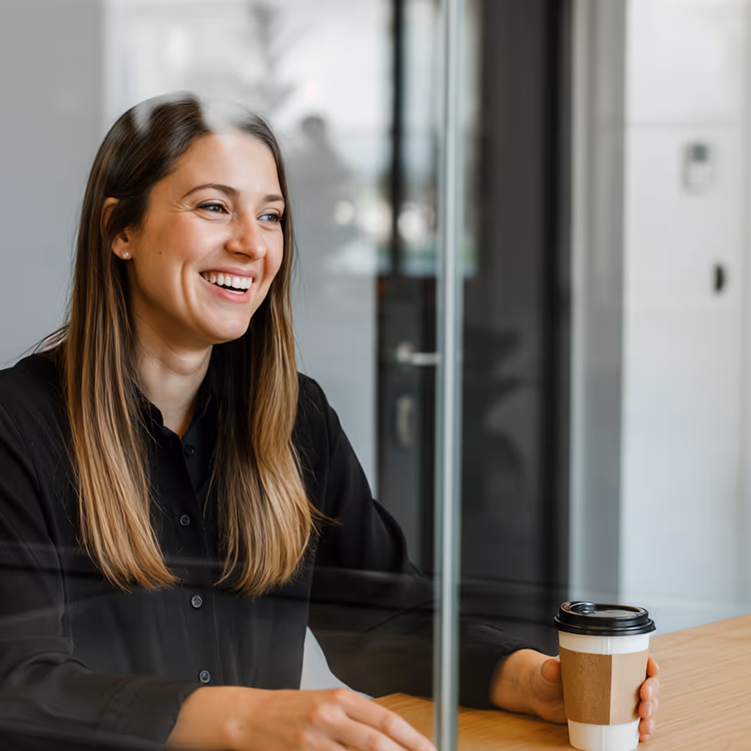 Leende kvinna i svart skjorta sitter vid ett bord och håller en papperskaffemugg med svart lock.