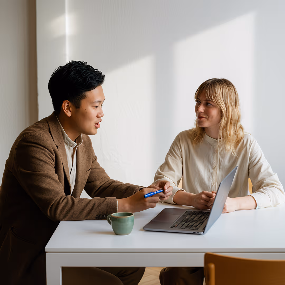 Två personer sitter vid ett bord och diskuterar med en öppen bärbar dator framför sig.