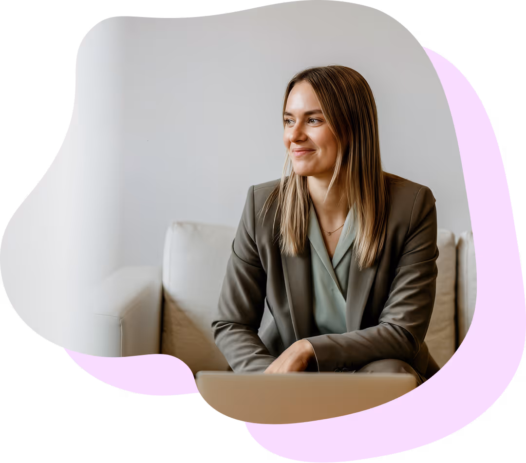 Kvinna med långt blont hår som sitter på en soffa och ler medan hon håller en bärbar dator.