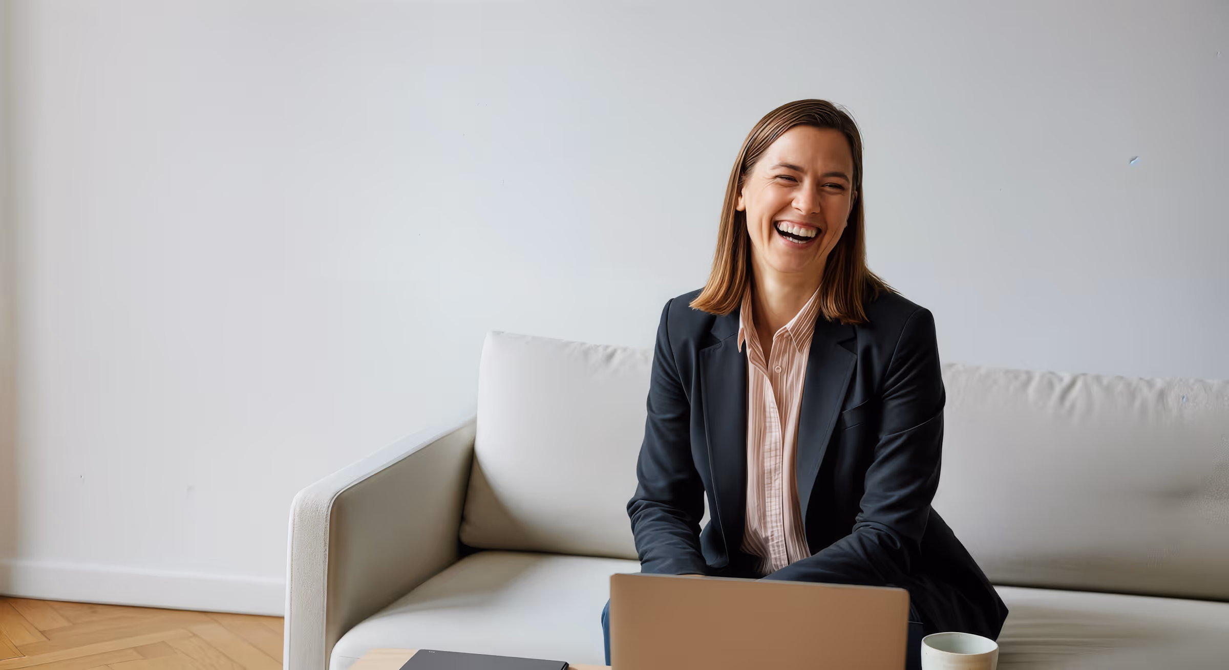 Leende kvinna i svart kavaj sitter på ljus soffa med laptop och mugg på bord framför.