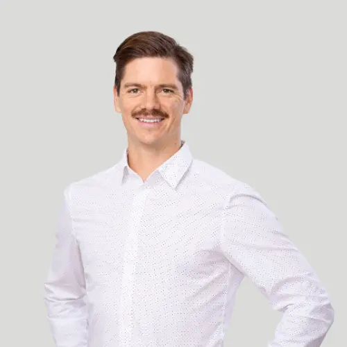 Man with mustache wearing a white button-up shirt standing against a light gray background.