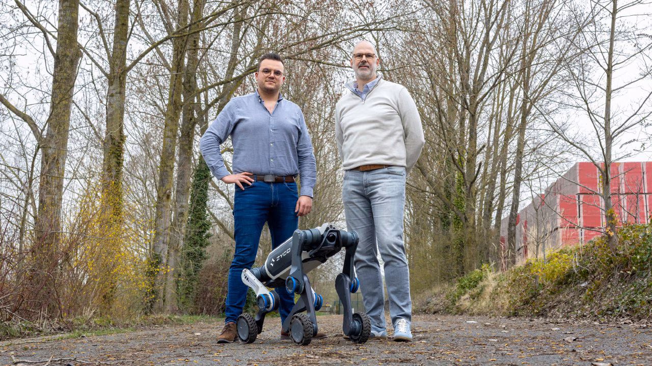 Two men standing outdoors on a path in front of a black four-legged robot dog with trees and buildings in the background.