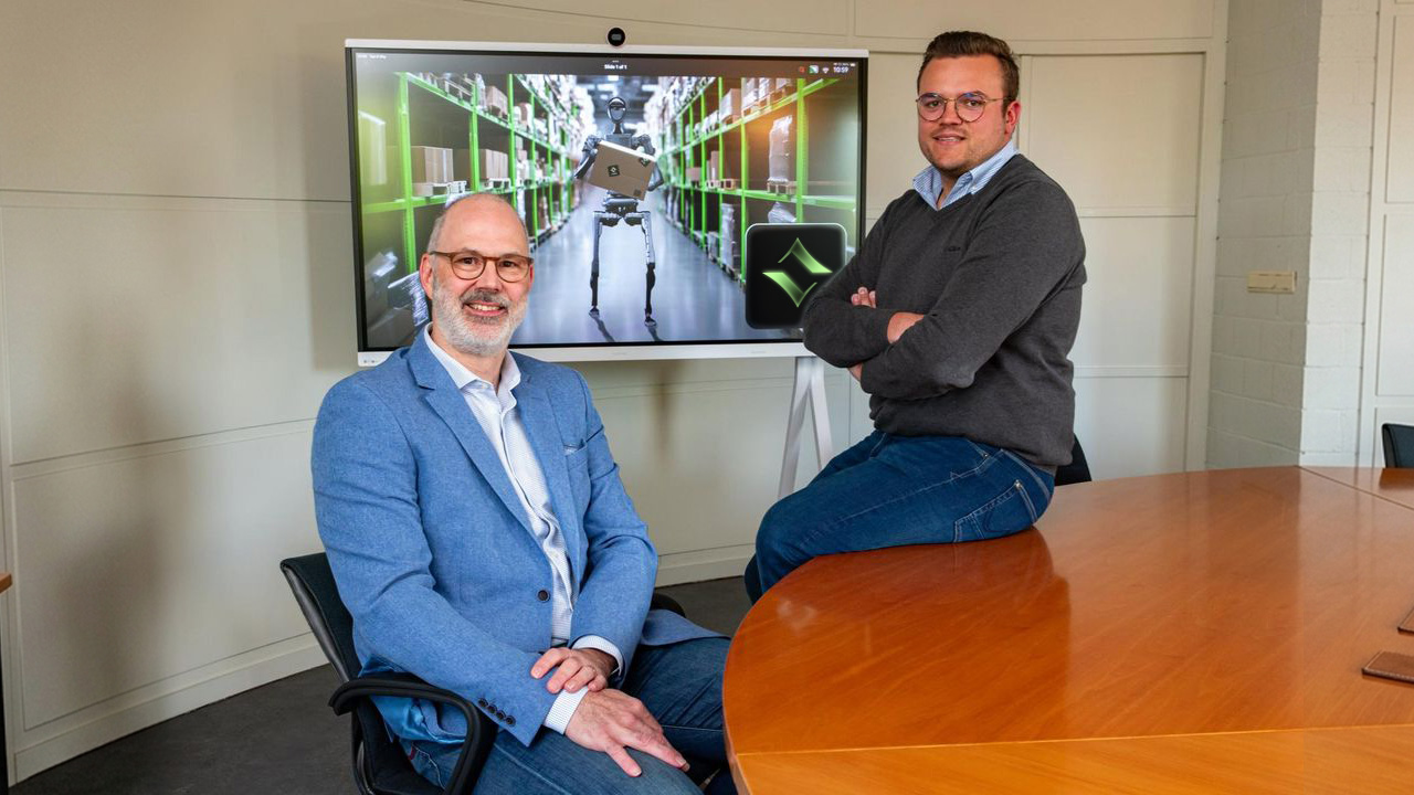 Two men sitting by a wooden table in an office with a screen behind them showing a robot carrying a box in a warehouse and the ZYGO logo.