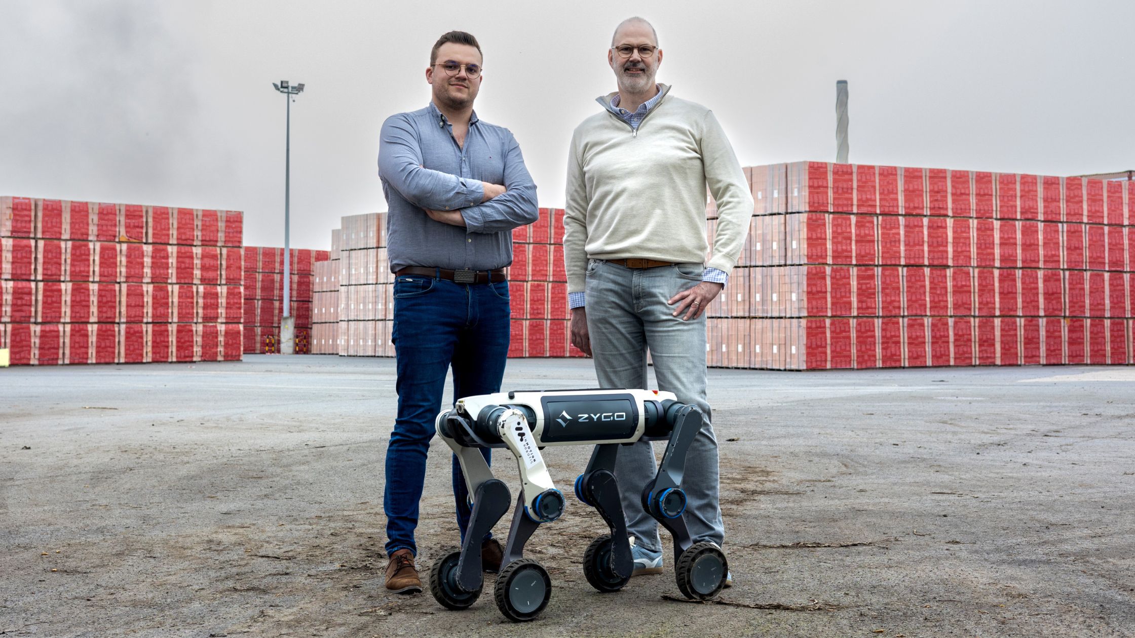 Two men standing outdoors on a path in front of a black four-legged robot dog with trees and buildings in the background.