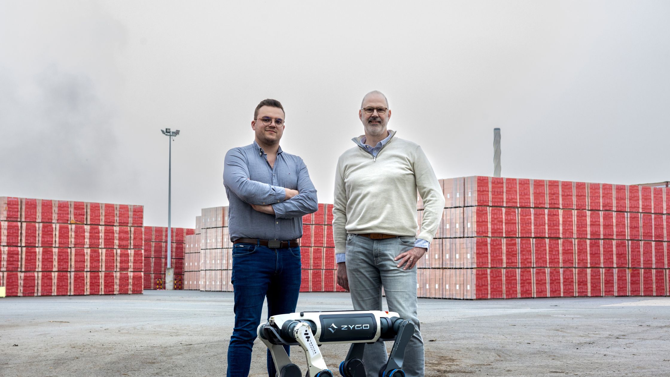 Two men standing outdoors on a path in front of a black four-legged robot dog with trees and buildings in the background.